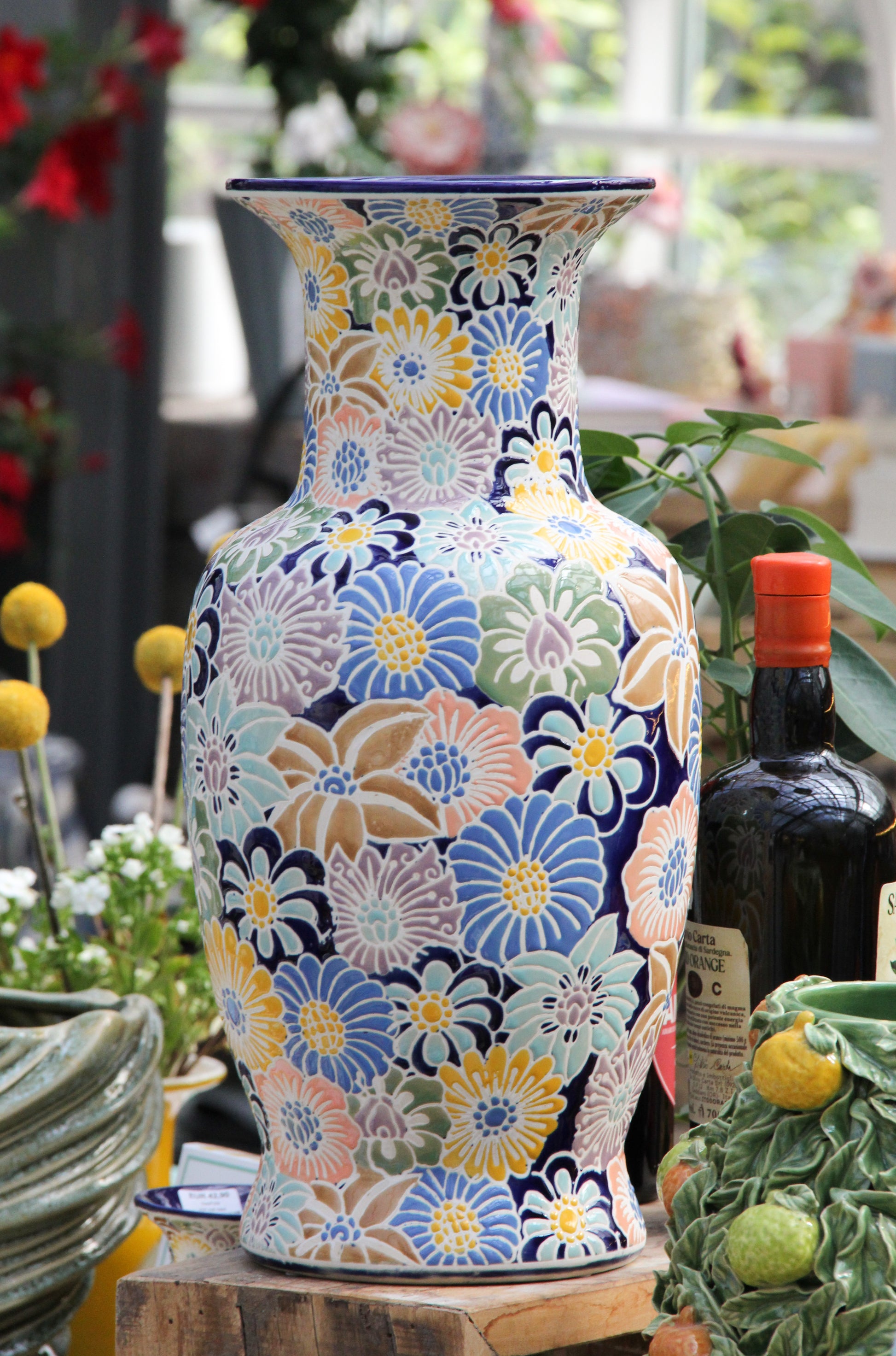 Eine bunte „Des Pots“-Vase aus glasierter Keramik mit Blumenmuster (H 40 cm, T 19,5 cm) steht auf einer Holzunterlage, umgeben von verschiedenen Pflanzen, einer Flasche Olivenöl und weiteren dekorativen Elementen in einem hellen Innenraum.
