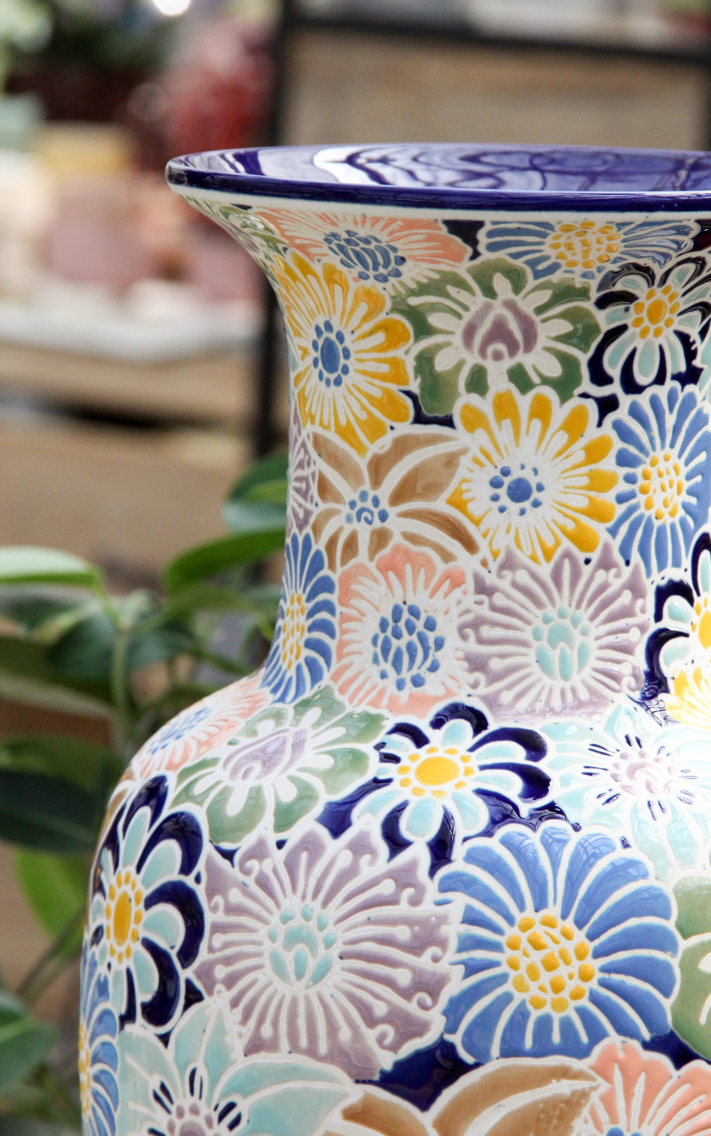 Eine Nahaufnahme der Vase „Des Pots | Bunte Vase aus Keramik mit Blumenmuster H 20 T 10“ von Des Pots, die sich durch farbenfrohe, handgemalte Blumenmuster in Blau-, Gelb-, Grün- und Pastelltönen vor einem verschwommenen Innenraumhintergrund auszeichnet. Diese glasierte Keramikvase ist 20 cm hoch und besticht durch exquisite Handwerkskunst.