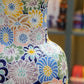 Eine Nahaufnahme der „Des Pots | Bunte Vase aus Keramik mit Blumenmuster“, einer glasierten Keramikvase von Des Pots mit breitem Rand und einer Höhe von 40 cm. Die Vase ist mit bunten Blumenmustern in Blau, Gelb, Grün, Pfirsich und Weiß verziert. Im unscharfen Hintergrund sind Regale mit verschiedenen Gegenständen zu sehen.