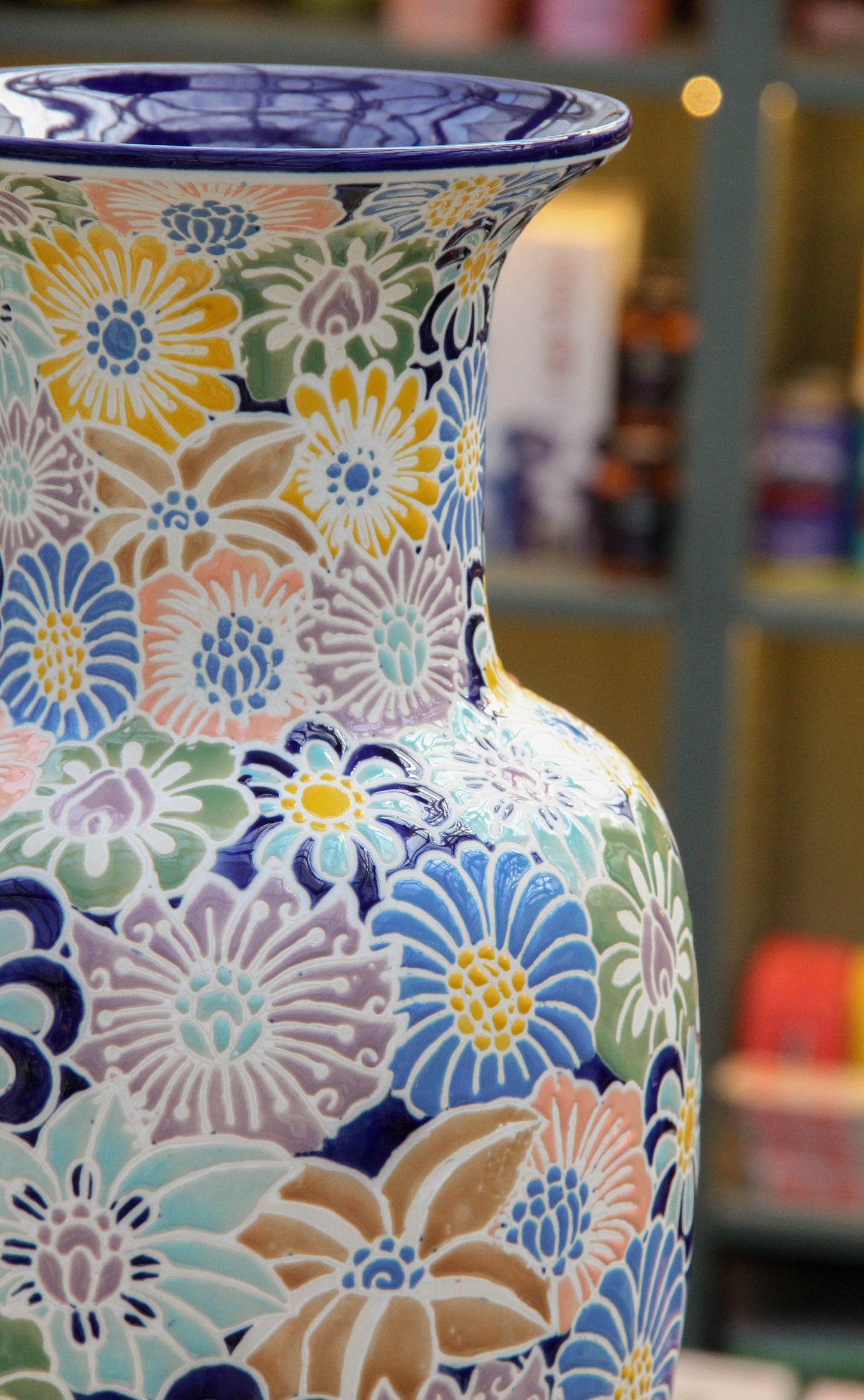 Eine Nahaufnahme der „Des Pots | Bunte Vase aus Keramik mit Blumenmuster“, einer glasierten Keramikvase von Des Pots mit breitem Rand und einer Höhe von 40 cm. Die Vase ist mit bunten Blumenmustern in Blau, Gelb, Grün, Pfirsich und Weiß verziert. Im unscharfen Hintergrund sind Regale mit verschiedenen Gegenständen zu sehen.