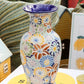 „A Des Pots | Bunte Vase aus Keramik mit Blumenmuster, H 20, T 10“ von Des Pots steht auf einem weißen Buch mit einem gelben Kreis und schwarzem Text. Im Hintergrund sind grüne Pflanzen und eine Karte mit Zitronen-Illustrationen zu sehen.