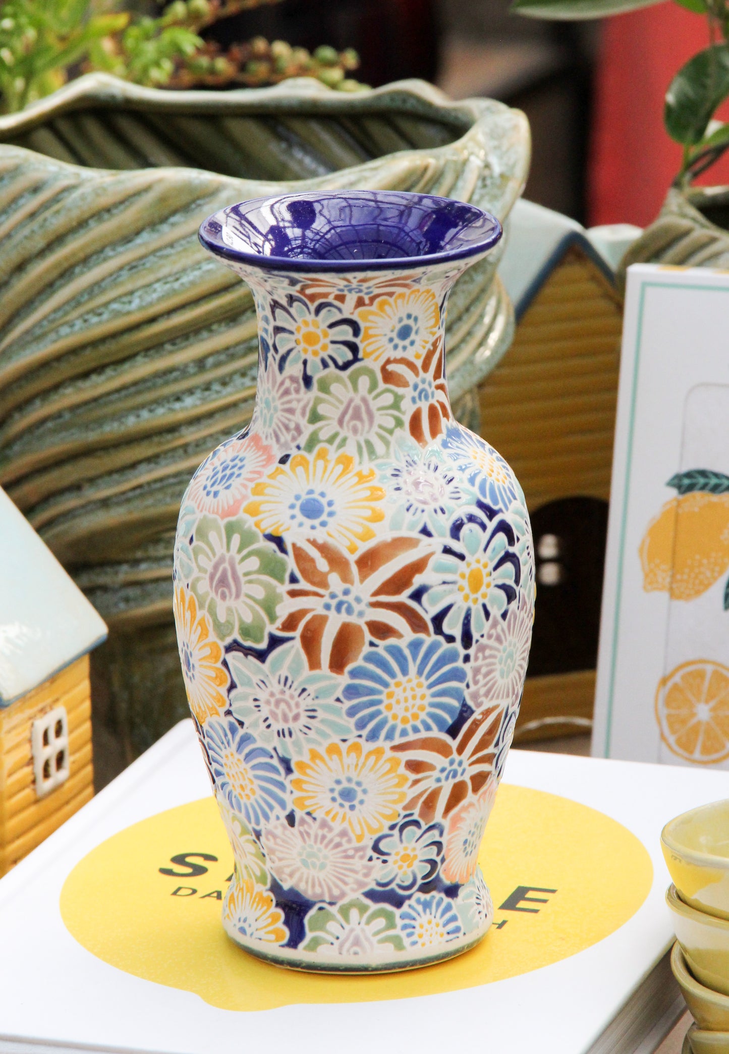 „A Des Pots | Bunte Vase aus Keramik mit Blumenmuster, H 20, T 10“ von Des Pots steht auf einem weißen Buch mit einem gelben Kreis und schwarzem Text. Im Hintergrund sind grüne Pflanzen und eine Karte mit Zitronen-Illustrationen zu sehen.