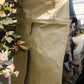 Eine Person, die die hellbraune Niwaki | Schürze „Split Apron“ (94 x 63 cm) und eine braune Hose trägt, steht neben einer üppig blühenden Pflanze. Aus ihrer Tasche ragt ein schwarzer Handschuh hervor, im Hintergrund sind verschwommene Gartenutensilien zu erkennen.