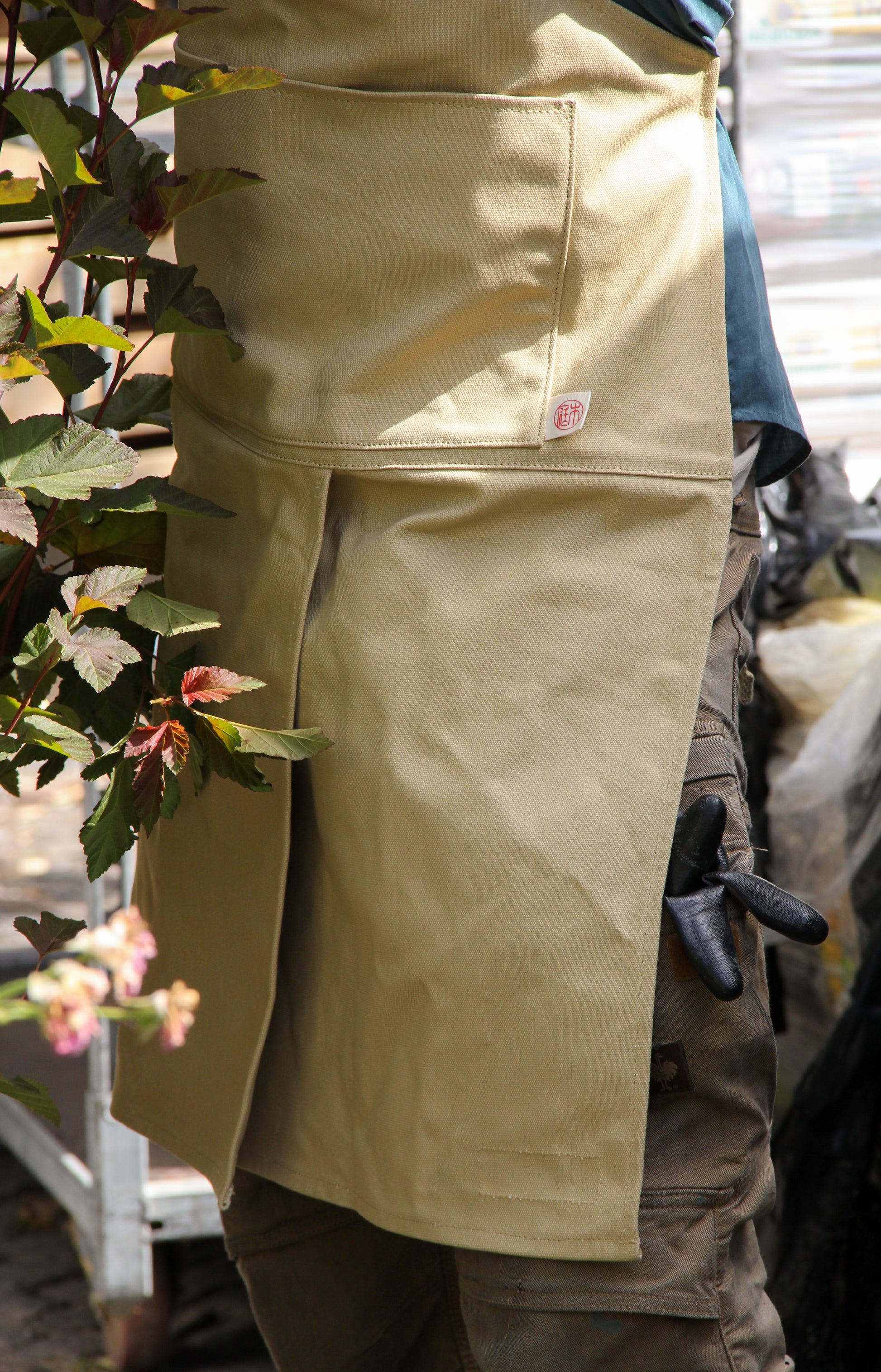 Eine Person, die die hellbraune Niwaki | Schürze „Split Apron“ (94 x 63 cm) und eine braune Hose trägt, steht neben einer üppig blühenden Pflanze. Aus ihrer Tasche ragt ein schwarzer Handschuh hervor, im Hintergrund sind verschwommene Gartenutensilien zu erkennen.