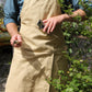 Eine Person, die eine hellbraune Niwaki | Schürze „Split Apron“ (94 x 63 cm) und ein blaues Hemd trägt, steht im Freien und hält ein kleines Handwerkzeug und ein Gartenmesser in der Hand, während sie neben grünen Blattpflanzen steht.