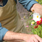 Eine Person, die eine hellbraune Niwaki-Schürze „Split Apron“ (94 x 63 cm) und ein blaues Hemd trägt, pflegt mit einer Gartenschere bunte Blumen in einem Garten. Zu sehen sind nur die Arme und ein Teil des Gesichts der Person.