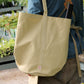 Eine Person in einem blauen Hemd und einer braunen Schürze steht im Freien und trägt die Niwaki Kantan Werkzeugtasche (beige, 26 cm hoch) von Niwaki über der Schulter. Im Hintergrund sind grüne Pflanzen und Holzregale zu sehen.