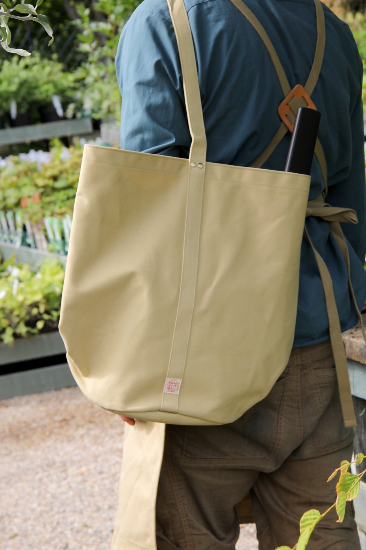 Eine Person in einem blauen Hemd und einer braunen Schürze steht im Freien und trägt die Niwaki Kantan Werkzeugtasche (beige, 26 cm hoch) von Niwaki über der Schulter. Im Hintergrund sind grüne Pflanzen und Holzregale zu sehen.
