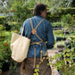Eine Person, die ein blaues Hemd und eine hellbraune Schürze trägt und die „Niwaki | Kantan Werkzeugtasche beige, 26 cm hoch“ von Niwaki bei sich hat, spaziert durch einen üppigen Garten, der von grünen Pflanzen und Laub umgeben ist.