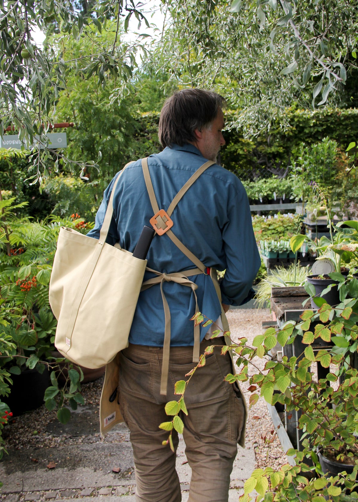 Eine Person, die ein blaues Hemd und eine hellbraune Schürze trägt und die „Niwaki | Kantan Werkzeugtasche beige, 26 cm hoch“ von Niwaki bei sich hat, spaziert durch einen üppigen Garten, der von grünen Pflanzen und Laub umgeben ist.