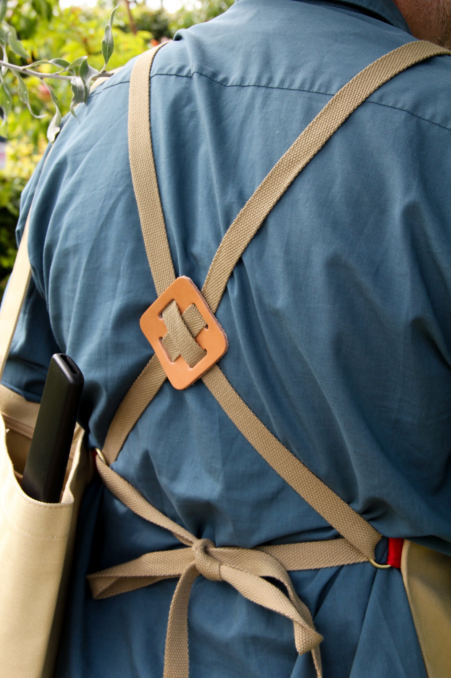Eine Person, die ein blaues Hemd und eine hellbraune Niwaki | Schürze „Split Apron“ (94 × 63 cm) trägt, ist von hinten zu sehen. Die Schürze verfügt über gekreuzte Träger mit einem Lederaufnäher in der Mitte, was für Niwakis hohen Qualitätsanspruch steht. In die Tasche der Schürze ist ein schwarzer Gegenstand gesteckt.