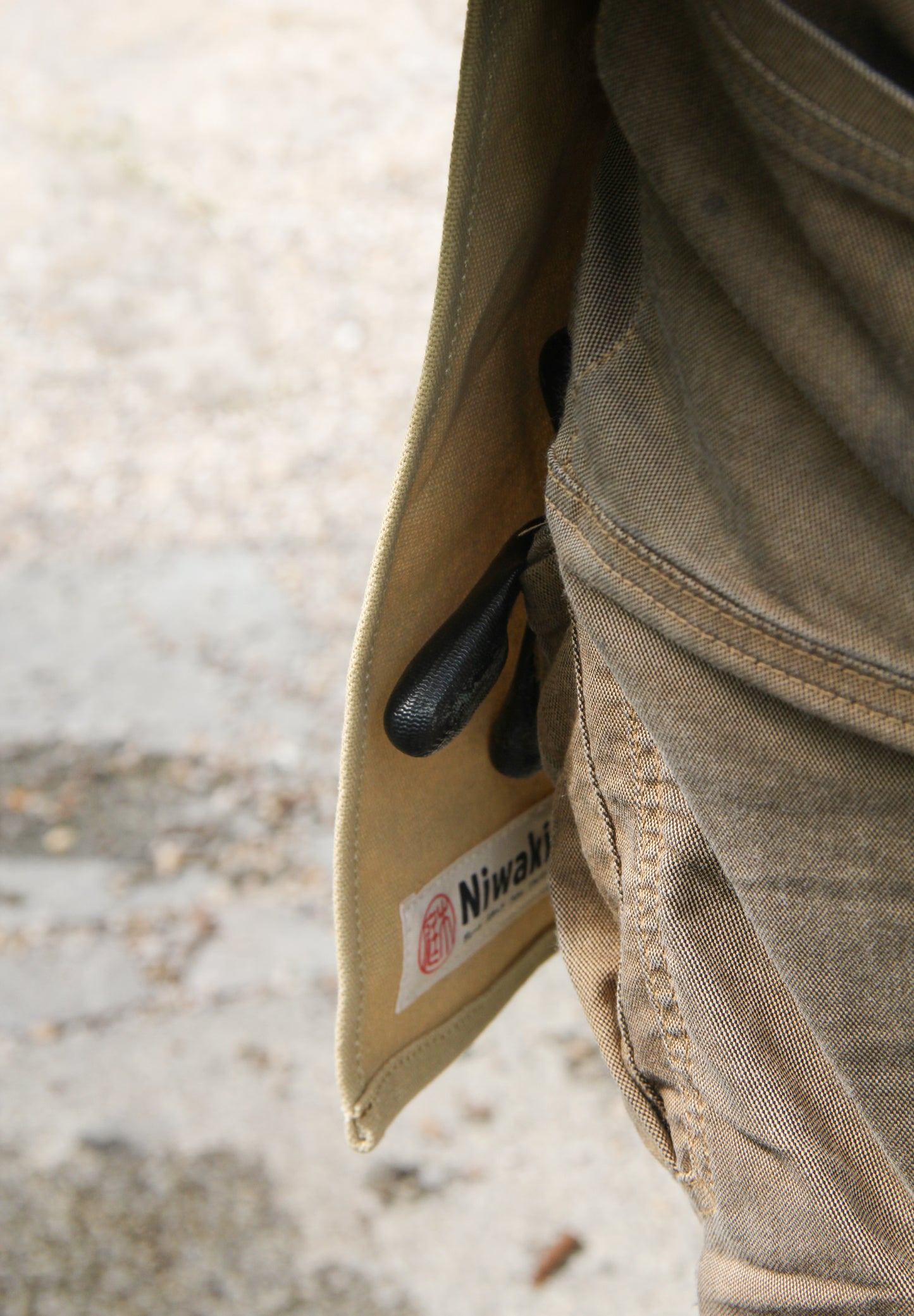 Eine Nahaufnahme einer Person, die eine braune Hose mit sichtbarem Niwaki-Etikett und einer schwarzen Gartenschere trägt, die teilweise in einer Tasche steckt, kombiniert mit der praktischen Niwaki | Schürze „Split Apron“ (94 x 63 cm) für mehr Funktionalität im Garten.