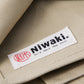 Eine Nahaufnahme des beigen Stoffes der Niwaki | Schürze „Split Apron“ (94 x 63 cm) zeigt ein weißes Etikett mit der Aufschrift „Niwaki“ in schwarzer Schrift und japanischen Schriftzeichen, ergänzt durch ein rotes, rundes Logo – ein Zeichen für die hochwertige Verarbeitung, die man von dieser Premium-Gartenschürze „Canvas“ von Niwaki erwartet.