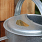 Nahaufnahme einer Zink-Gießkanne „Plint | Gießkanne klein 1,5 l“ von PLINT A/S mit geschwungenem Henkel, die neben einer Holzfläche steht. Auf einem goldenen Etikett auf der Gießkanne steht „PLANT“.