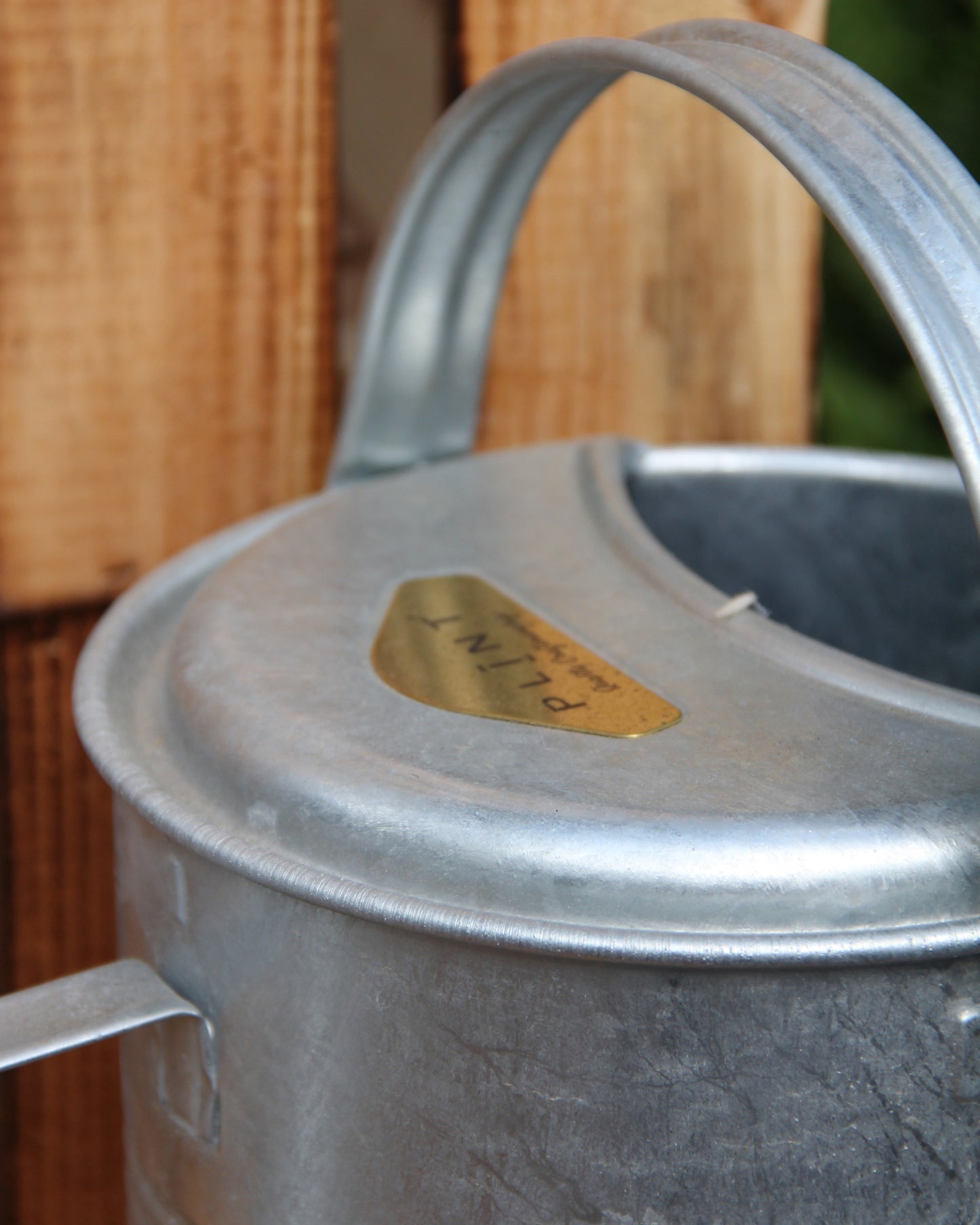 Nahaufnahme einer Zink-Gießkanne „Plint | Gießkanne klein 1,5 l“ von PLINT A/S mit geschwungenem Henkel, die neben einer Holzfläche steht. Auf einem goldenen Etikett auf der Gießkanne steht „PLANT“.