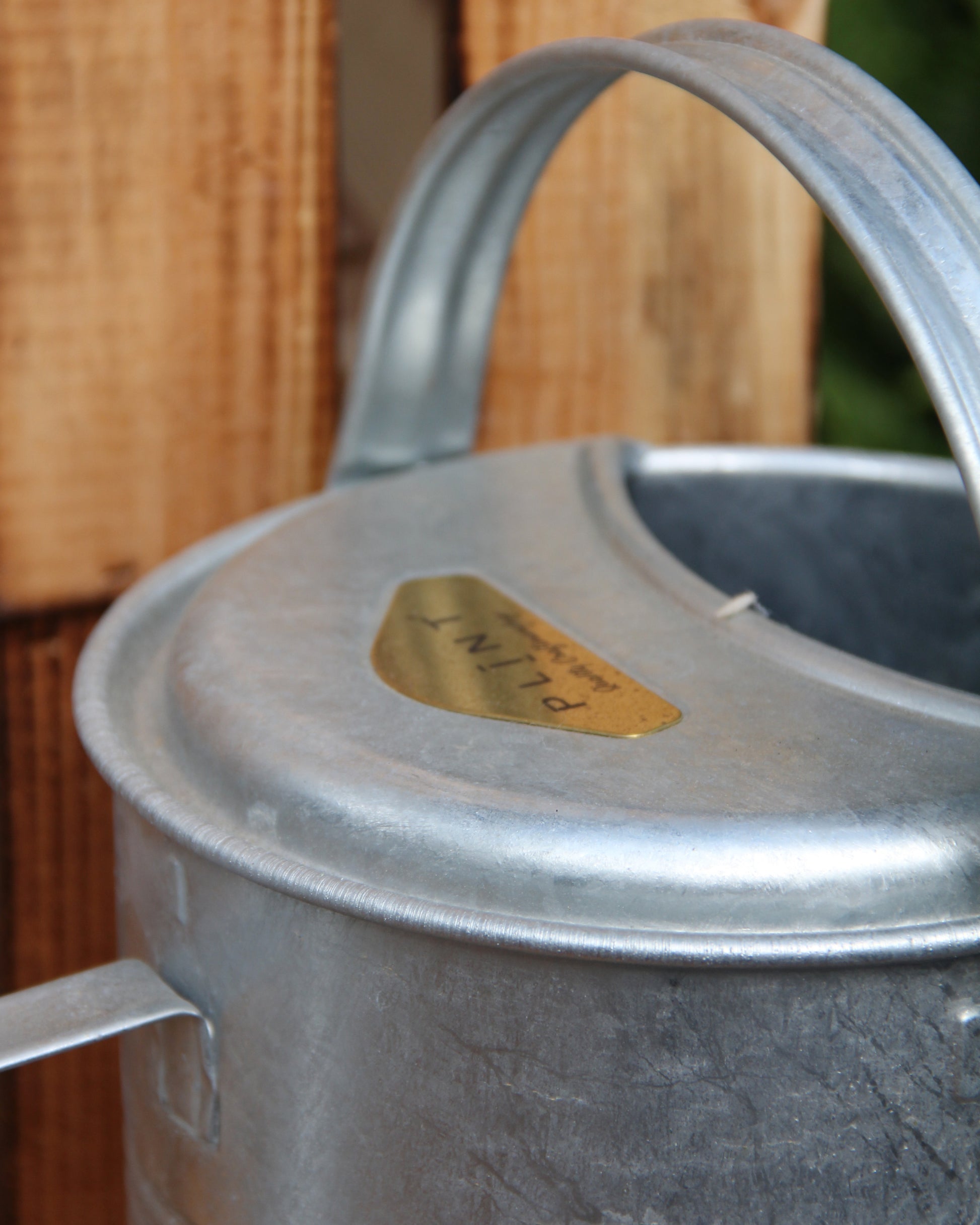 Nahaufnahme einer Zink-Gießkanne „Plint | Gießkanne klein 1,5 l“ von PLINT A/S mit geschwungenem Henkel, die neben einer Holzfläche steht. Auf einem goldenen Etikett auf der Gießkanne steht „PLANT“.