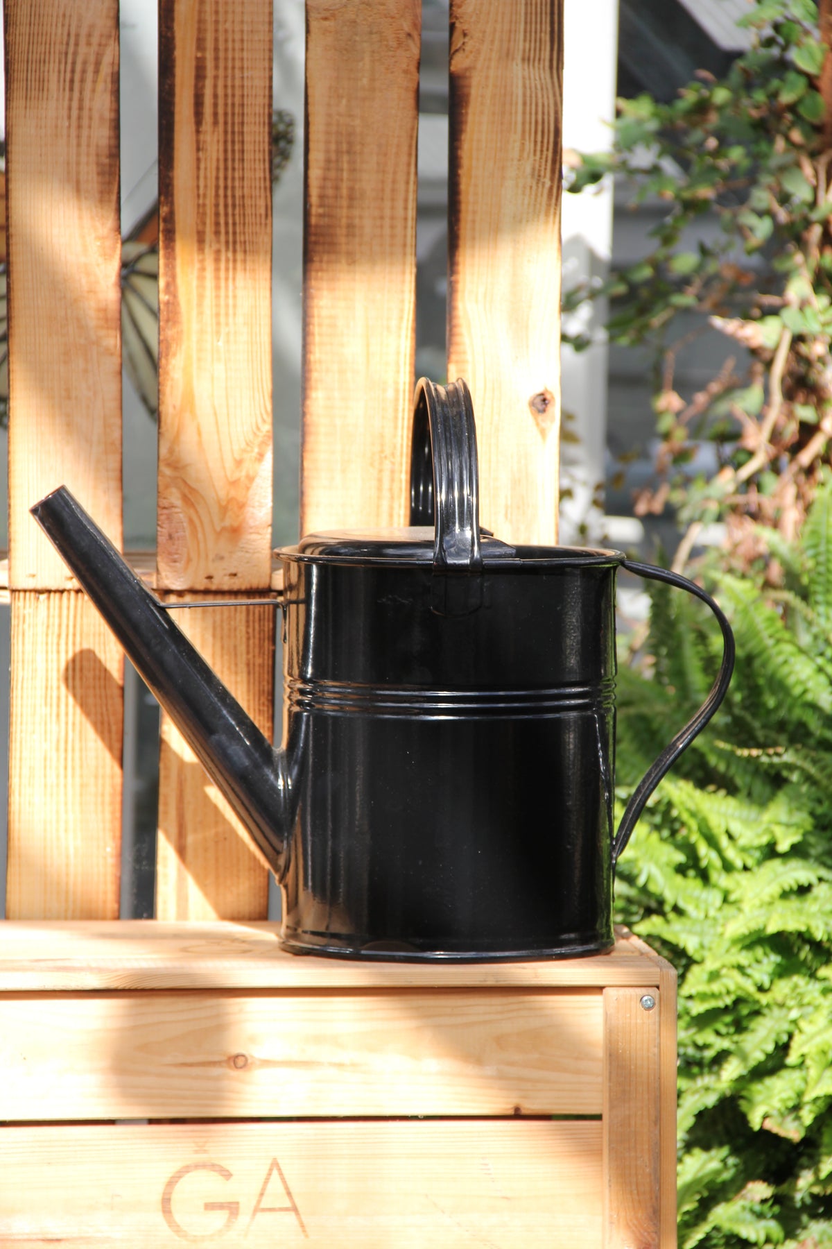 Eine „Plint | Gießkanne klein 1,5 l in Schwarz“ von PLINT A/S steht auf einer Holzbank im Freien, während das Sonnenlicht Schatten wirft. Im Hintergrund sind grüne Farne und eine Holzpalette zu sehen.