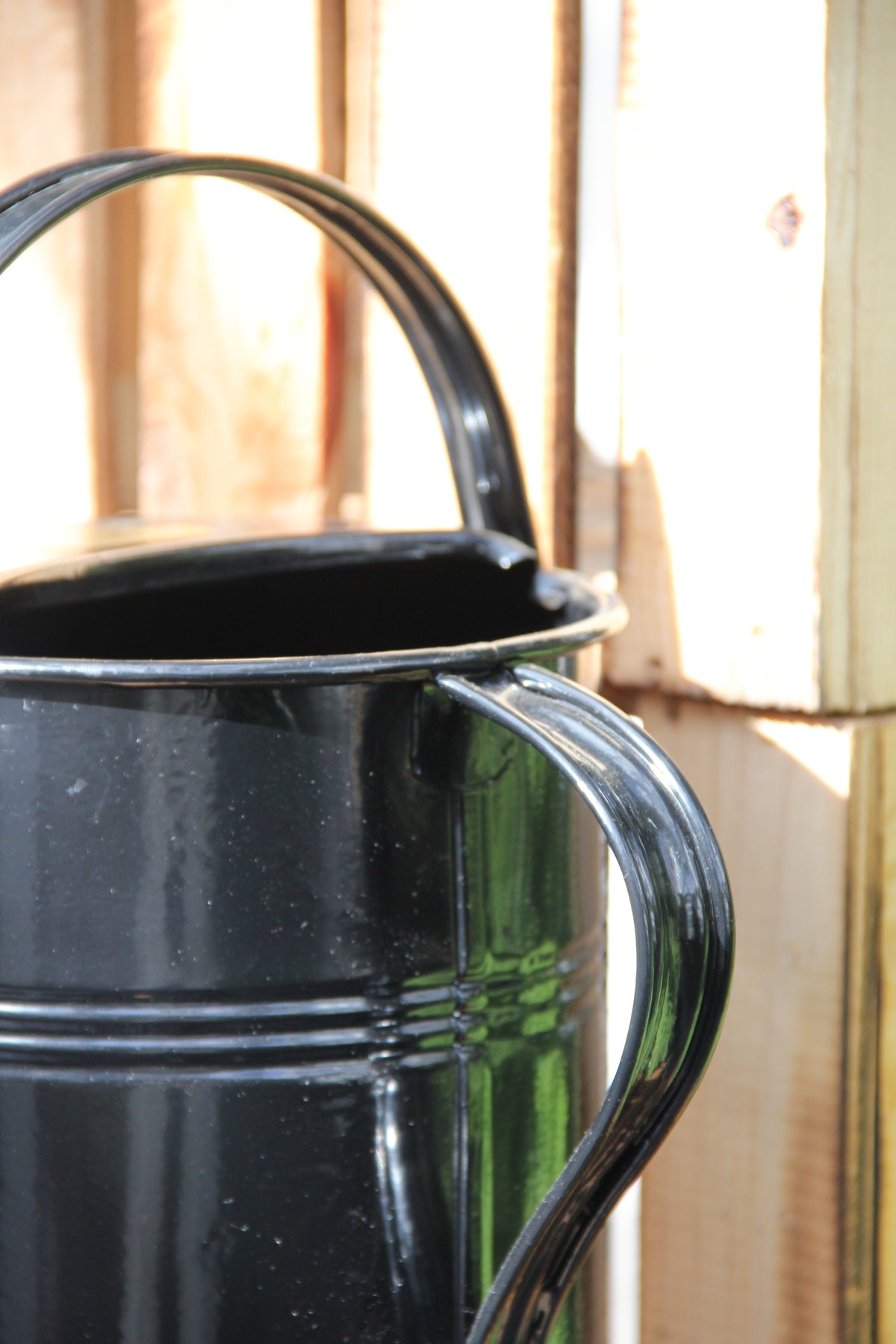 Eine Nahaufnahme einer glänzend schwarzen „Plint | Gießkanne klein 1,5 L in Schwarz“ von PLINT A/S mit zwei Griffen, platziert vor einem Holzhintergrund, auf dem das Sonnenlicht Schatten wirft.