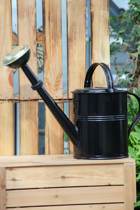 Eine Gießkanne „Plint“ | Gießkanne klein 1,5 l in Grün von PLINT A/S mit langem Auslauf steht auf einer Holzbank in einem Garten, im Hintergrund sind ein Holzzaun und grüne Pflanzen zu sehen.