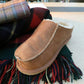 Ein Richard Slipper aus Schaffell in Cognac (Größe 42) von Shepherd of Sweden liegt gemütlich auf einem gewollten Schal mit roten, grünen und marineblauen Streifen; im Hintergrund befindet sich eine zusammengerollte Tartan-Decke.