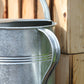 Eine Nahaufnahme der „Plint | Gießkanne Medium 5 l in Zink“ von PLINT A/S mit geschwungenem Henkel, platziert neben einer Holzwand. Das Sonnenlicht bringt die Zinkoberfläche und die Holzmaserung besonders gut zur Geltung und unterstreicht die hohe Qualität dieser 5-Liter-Gießkanne.