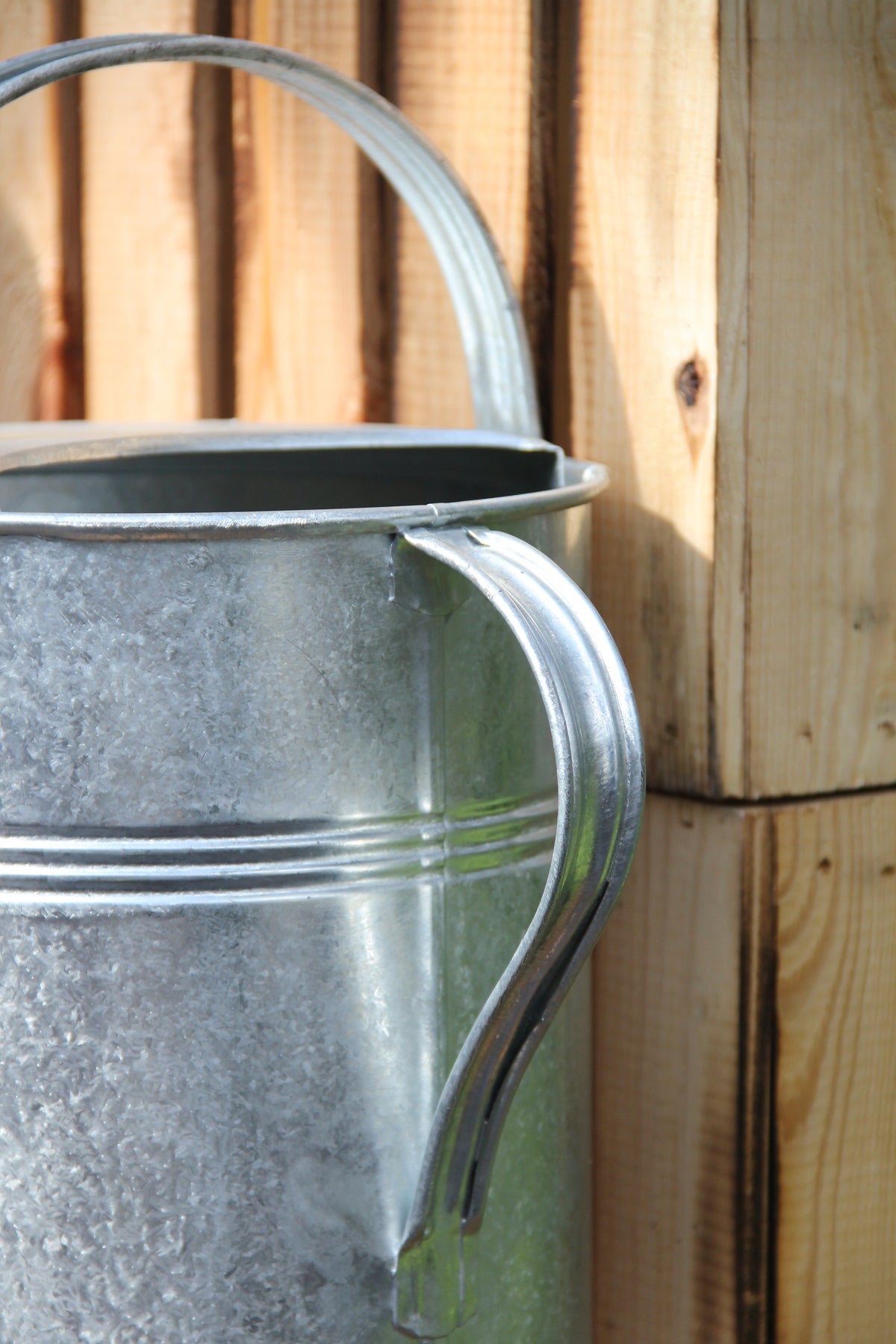 Eine Nahaufnahme der „Plint | Gießkanne Medium 5 l in Zink“ von PLINT A/S mit geschwungenem Henkel, platziert neben einer Holzwand. Das Sonnenlicht bringt die Zinkoberfläche und die Holzmaserung besonders gut zur Geltung und unterstreicht die hohe Qualität dieser 5-Liter-Gießkanne.