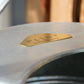Nahaufnahme einer Gießkanne „Medium“ (5 l) von PLINT A/S aus Zink mit einem goldenen Etikett, auf dem „Plint Quality Craftsmanship“ steht, vor dem Hintergrund verschwommener Holzbretter.