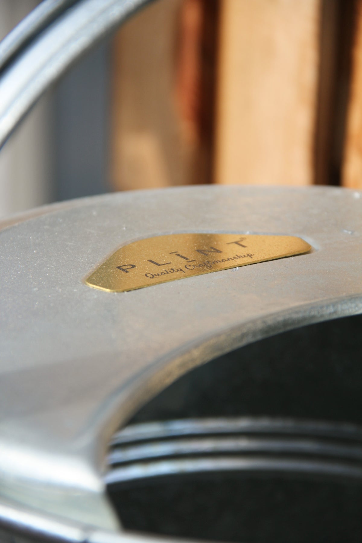 Nahaufnahme einer Gießkanne „Medium“ (5 l) von PLINT A/S aus Zink mit einem goldenen Etikett, auf dem „Plint Quality Craftsmanship“ steht, vor dem Hintergrund verschwommener Holzbretter.