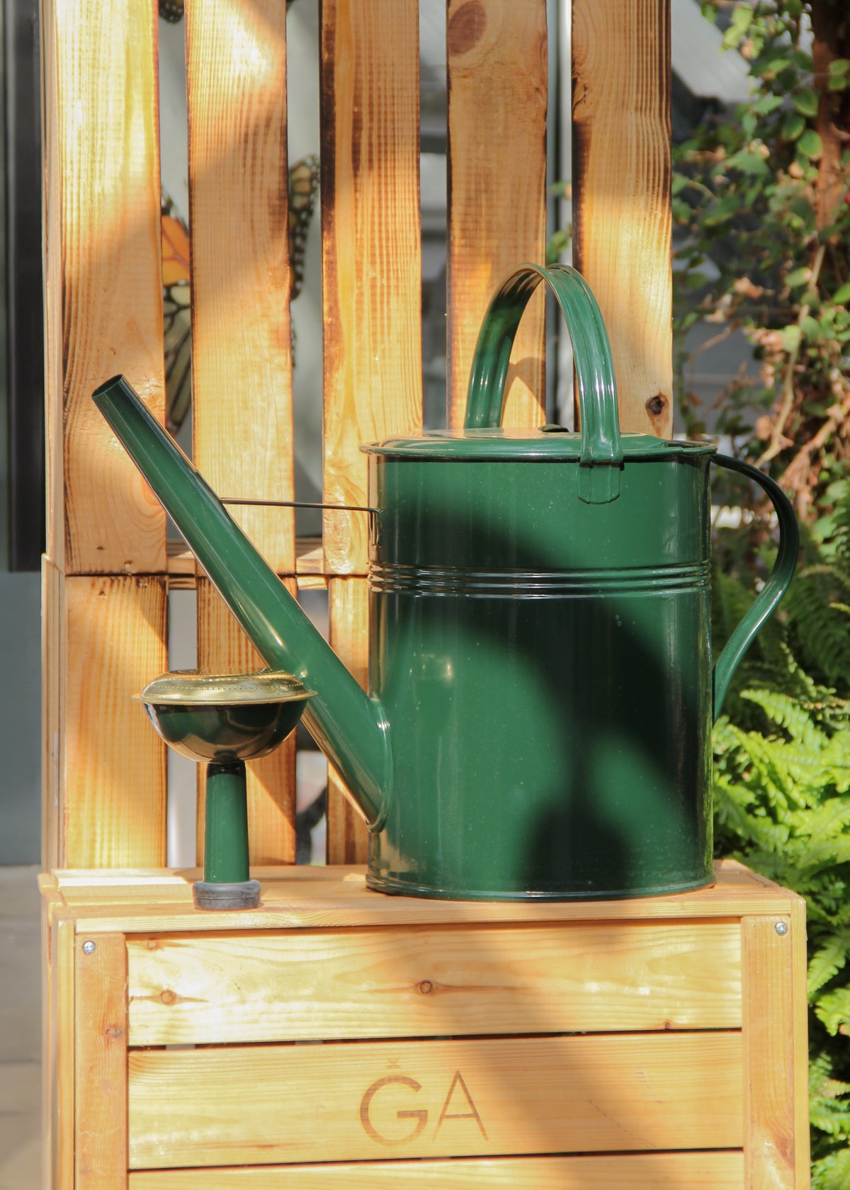 Eine glänzende, dunkelgrüne „Plint | Gießkanne Medium 5 l“ von PLINT A/S aus verzinktem Stahl steht zusammen mit einem kleinen, dazu passenden Becher auf einer Holzkiste, in die „GA“ eingraviert ist. Das Arrangement liegt im Sonnenlicht, vor dem Hintergrund üppiger Pflanzen und einer Holzpalette.