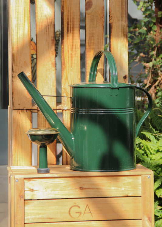 Eine glänzende, dunkelgrüne „Plint | Gießkanne Medium 5 l“ von PLINT A/S aus verzinktem Stahl steht zusammen mit einem kleinen, dazu passenden Becher auf einer Holzkiste, in die „GA“ eingraviert ist. Das Arrangement liegt im Sonnenlicht, vor dem Hintergrund üppiger Pflanzen und einer Holzpalette.