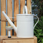 Eine Gießkanne „Plint“ Medium 9 l in Weiß von PLINT A/S steht auf einer Holzkiste neben einem kleinen Gartenwasserhahn, mit einem Holzzaun und üppigen grünen Pflanzen im Hintergrund – ideal für eine effiziente Pflanzenbewässerung.