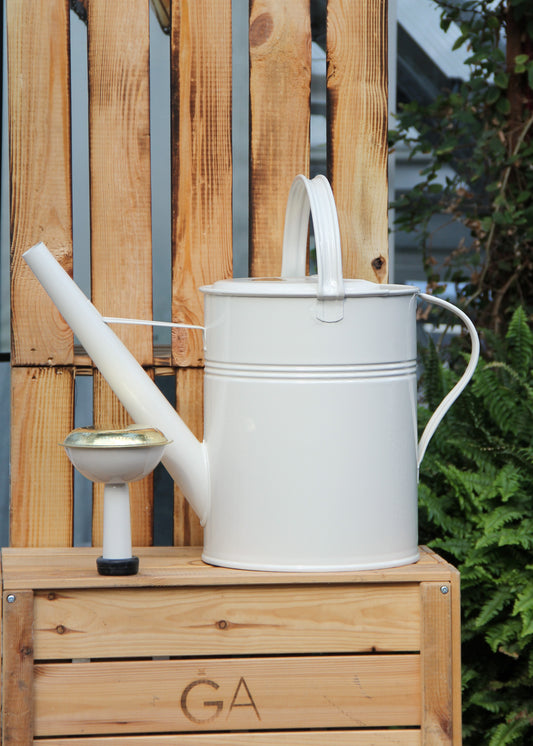 Eine Gießkanne „Plint“ Medium 9 l in Weiß von PLINT A/S steht auf einer Holzkiste neben einem kleinen Gartenwasserhahn, mit einem Holzzaun und üppigen grünen Pflanzen im Hintergrund – ideal für eine effiziente Pflanzenbewässerung.