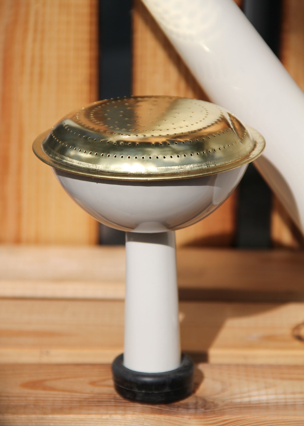 Eine Nahaufnahme des Auslaufs der „Plint | Gießkanne Medium 9 l in Weiß“ von PLINT A/S, mit einer runden, goldfarbenen, perforierten Rosette vor einem Holzhintergrund – perfekt für eine effektive Pflanzenbewässerung.
