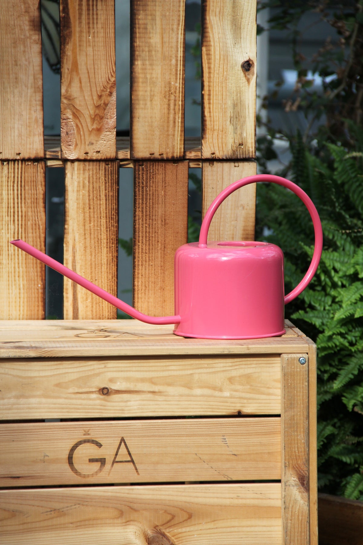Eine Gießkanne aus Metall | 1 l in leuchtendem Pink von Rex Europe mit langem Ausguss steht auf einer mit „GA“ beschrifteten Holzkiste vor einer Holzpalette, im Hintergrund sind grüne Pflanzen zu sehen. Diese stilvolle Gießkanne vereint Charme und Funktionalität mit ihrem Fassungsvermögen von 1 Liter und eignet sich somit perfekt für die Gartenpflege.