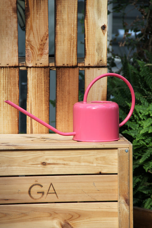 Eine Gießkanne aus Metall | 1 l in leuchtendem Pink von Rex Europe mit langem Ausguss steht auf einer mit „GA“ beschrifteten Holzkiste vor einer Holzpalette, im Hintergrund sind grüne Pflanzen zu sehen. Diese stilvolle Gießkanne vereint Charme und Funktionalität mit ihrem Fassungsvermögen von 1 Liter und eignet sich somit perfekt für die Gartenpflege.