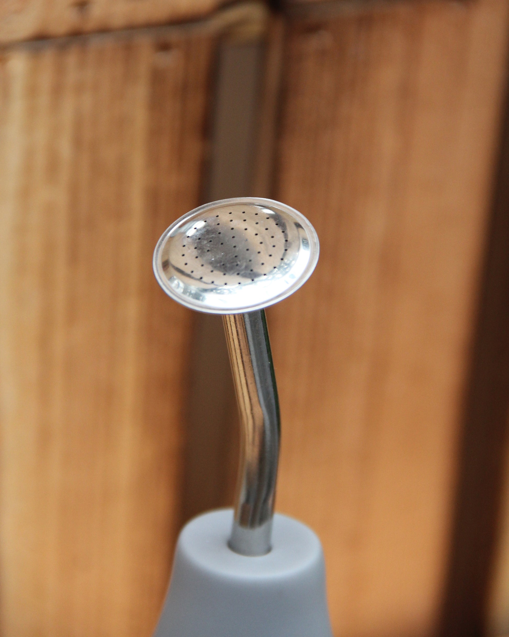 Eine Nahaufnahme der „Esschert Gießbrause Ball Grau L“, einer Gießkanne aus Metall mit kleinen Löchern für eine wassersparende und gezielte Pflanzenbewässerung, die an einem weißen Griff befestigt ist und vor einem Holzhintergrund steht.