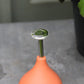 Eine „Esschert Gießbrause Ball Terracotta S“, eine kompakte, terrakotta-orangefarbene Gießbrause für die gezielte und wassersparende Pflanzenpflege, steht auf einer grauen Fläche neben grünen Pflanzen.