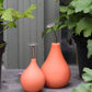 Zwei Esschert-Gießkannen vom Typ „Gießbrause Ball Terracotta S“ mit orangefarbenen, knollenförmigen Körpern und silbernen Ausgüssen stehen auf einer grauen Fläche inmitten grüner Topfpflanzen.