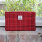 Eine handgewebte Tasche mit Reißverschluss | „Harris Tweed Berneray“ Medium Red Check 23 x 11 cm von Glen Appin, mit einem klassischen roten Karomuster und einem kleinen Etikett, liegt auf einem ziegelroten Fensterbrett. Hinter ihr sind durch das Fenster grüne Blätter zu sehen.