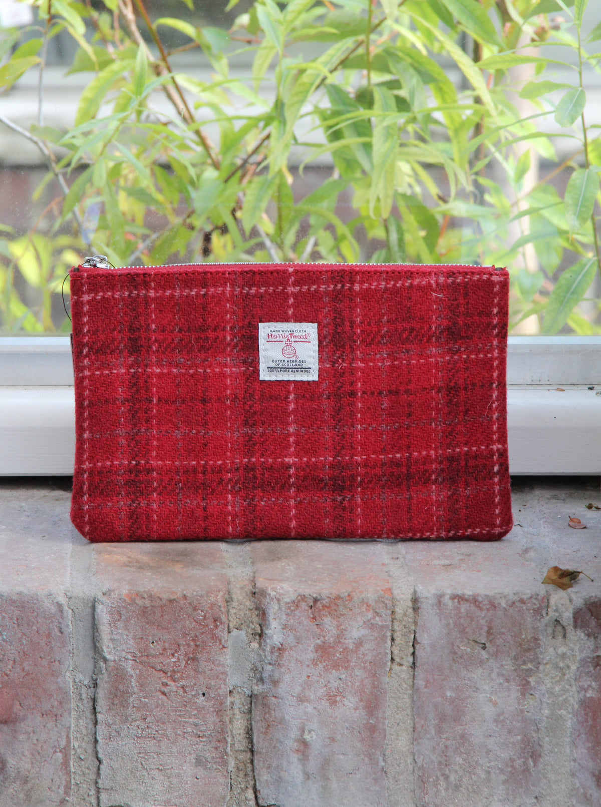 Eine handgewebte Tasche mit Reißverschluss | „Harris Tweed Berneray“ Medium Red Check 23 x 11 cm von Glen Appin, mit einem klassischen roten Karomuster und einem kleinen Etikett, liegt auf einem ziegelroten Fensterbrett. Hinter ihr sind durch das Fenster grüne Blätter zu sehen.