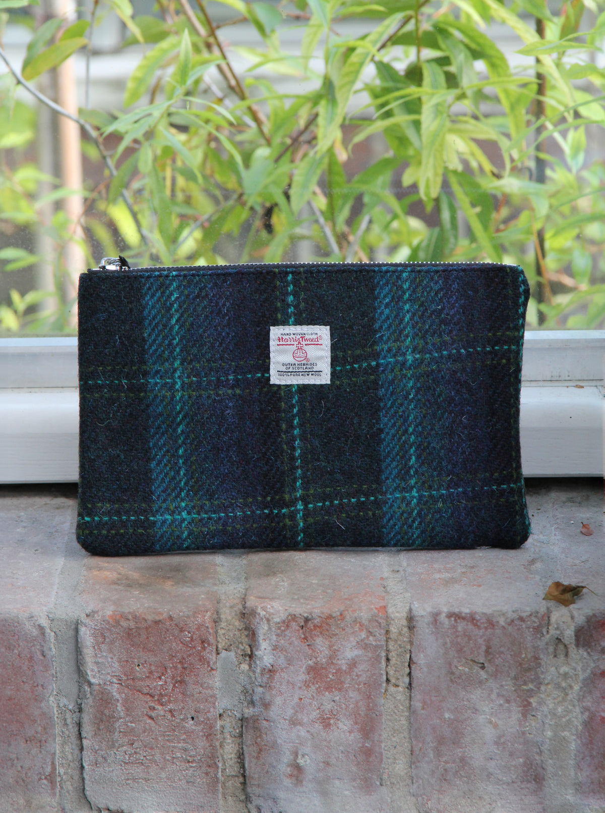 Eine kleine Tasche mit Reißverschluss, die „Glen Appin Handgewebte Tasche mit Reißverschluss | ‚Berneray‘ Medium“ in Türkisblau mit türkisfarbenem Karomuster (23 x 11 cm), liegt auf einem gemauerten Fensterbrett, während draußen vor dem Fenster grüne Blätter zu sehen sind.