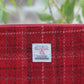 Eine Nahaufnahme der „Harris Tweed Berneray“ – einer handgewebten Tasche mit Reißverschluss von Glen Appin – zeigt den klassischen Wollstoff „Medium Red Check“ und das aufgenähte, ikonische Harris-Tweed-Etikett. Die Detailaufnahme unterstreicht die handwerkliche Qualität dieses 23 x 11 cm großen, handgewebten Accessoires, wobei ein sanft unscharfer Hintergrund aus grünen Blättern dessen handwerkliche Qualität besonders hervorhebt.