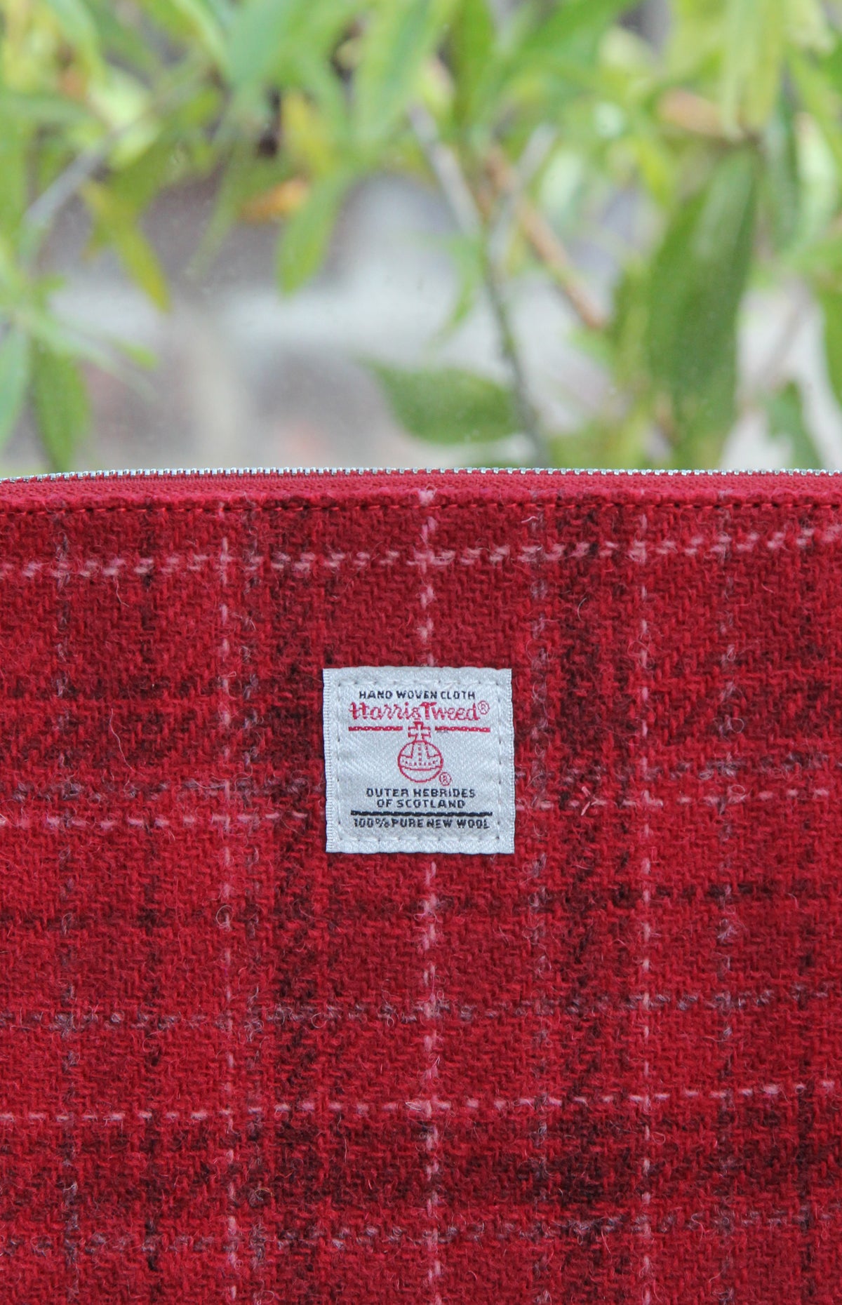 Eine Nahaufnahme der „Harris Tweed Berneray“ – einer handgewebten Tasche mit Reißverschluss von Glen Appin – zeigt den klassischen Wollstoff „Medium Red Check“ und das aufgenähte, ikonische Harris-Tweed-Etikett. Die Detailaufnahme unterstreicht die handwerkliche Qualität dieses 23 x 11 cm großen, handgewebten Accessoires, wobei ein sanft unscharfer Hintergrund aus grünen Blättern dessen handwerkliche Qualität besonders hervorhebt.