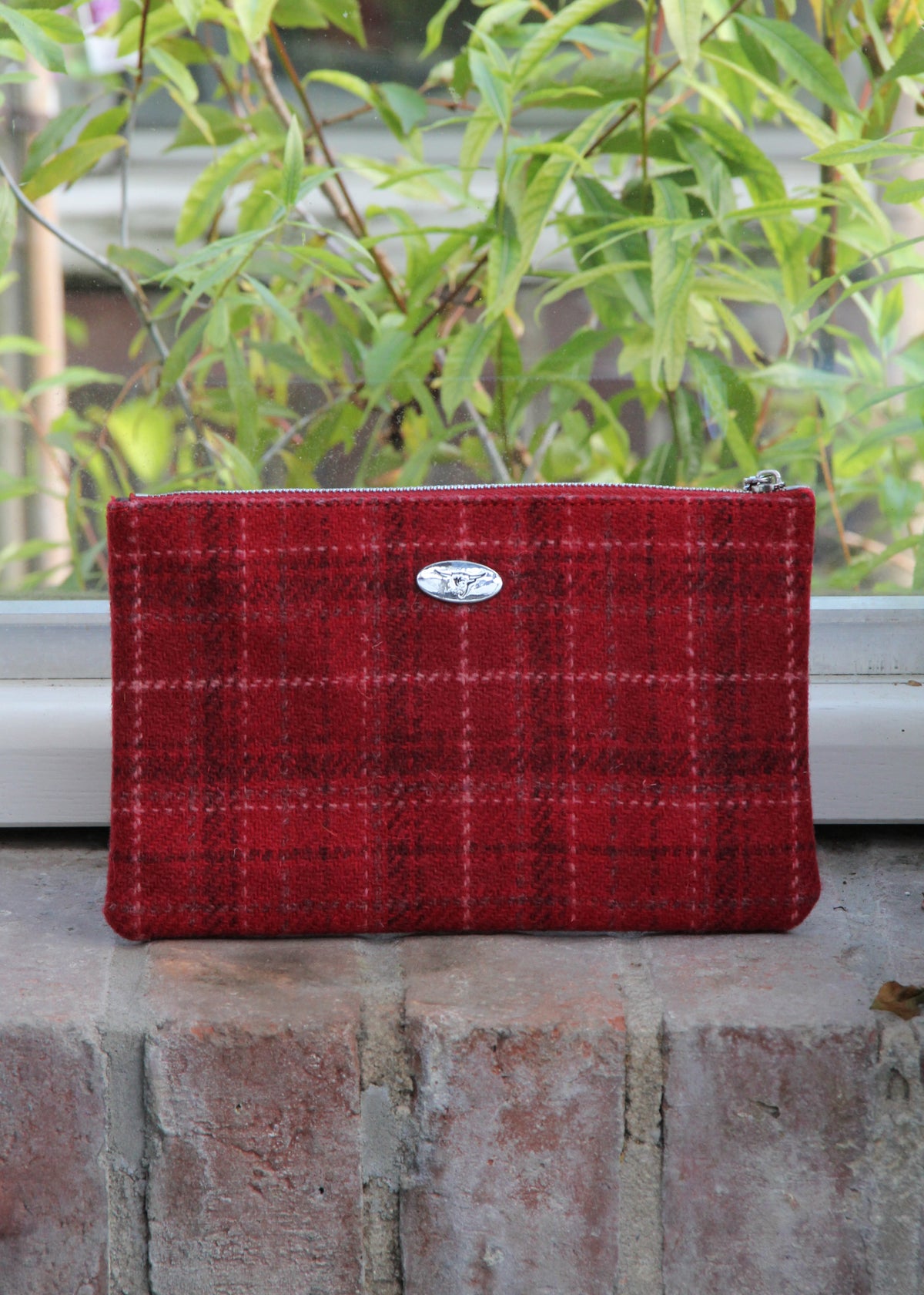 Eine handgewebte Tasche aus „Harris Tweed Berneray“ mit mittelrotem Karomuster und Reißverschluss (23 x 11 cm) von Glen Appin, verziert mit einem silbernen, ovalen Emblem, liegt auf einem Backsteinfenstersims vor einem Fenster, vor dem draußen grüne Blattpflanzen stehen.