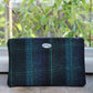 Eine handgewebte Tasche mit Reißverschluss aus der Kollektion von Glen Appin, Modell „Berneray“ in Medium Turquoise Blue mit türkisfarbenem Überkaro (23 x 11 cm), liegt auf einem Backsteinvorsprung vor grünem Laub im Hintergrund.