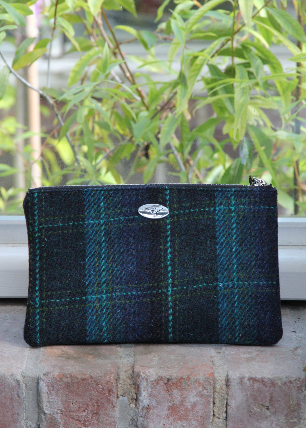Eine handgewebte Tasche mit Reißverschluss aus der Kollektion von Glen Appin, Modell „Berneray“ in Medium Turquoise Blue mit türkisfarbenem Überkaro (23 x 11 cm), liegt auf einem Backsteinvorsprung vor grünem Laub im Hintergrund.