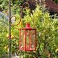 In einem sonnigen Garten hängt eine rote Metalllaterne am Hirsch-Laternenstab „Bino Pinie“, umgeben von einem Orangenbaum mit reifenden Früchten.