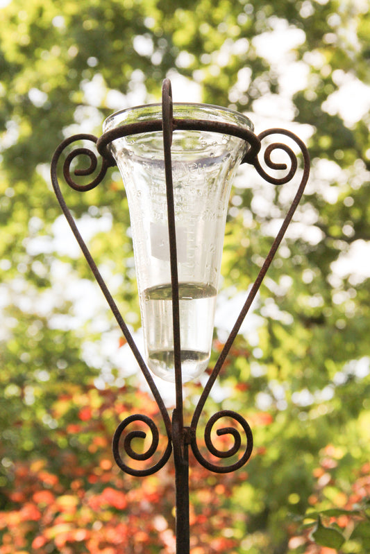 Ein Regenmesser mit Glas | Für den Gartenliebhaber, inkl. filigranem Universalstab, H 125 cm, von Hirsch, teilweise mit Wasser gefüllt, steht aufrecht in seinem dekorativen Metallständer, im Hintergrund ist sanft verschwommenes grünes und rotes Laub zu sehen.