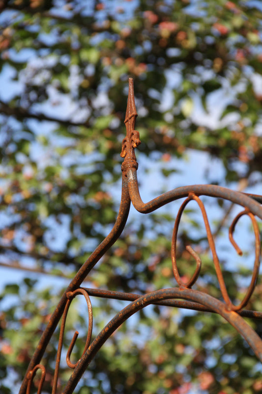 Eine Nahaufnahme eines feuerverzinkten Metallzauns mit dem Modell „Hirsch Rosenbogen SPITZ Vollmetall stabil“ aus massivem 12-mm-Material vor einem verschwommenen Hintergrund aus grünen Zweigen und blauem Himmel.