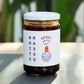 Ein 100-ml-Glas White Mausu | Rauchiges Chiliöl von White Mausu Ltd. ruht auf einer grauen Oberfläche, mit verschwommenen grünen Pflanzen im Hintergrund, die den rauchigen Geschmack hervorheben.